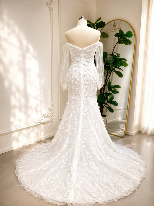 White mermaid wedding dress with 3D floral appliqués, off-the-shoulder illusion sleeves, and a long flowing train.