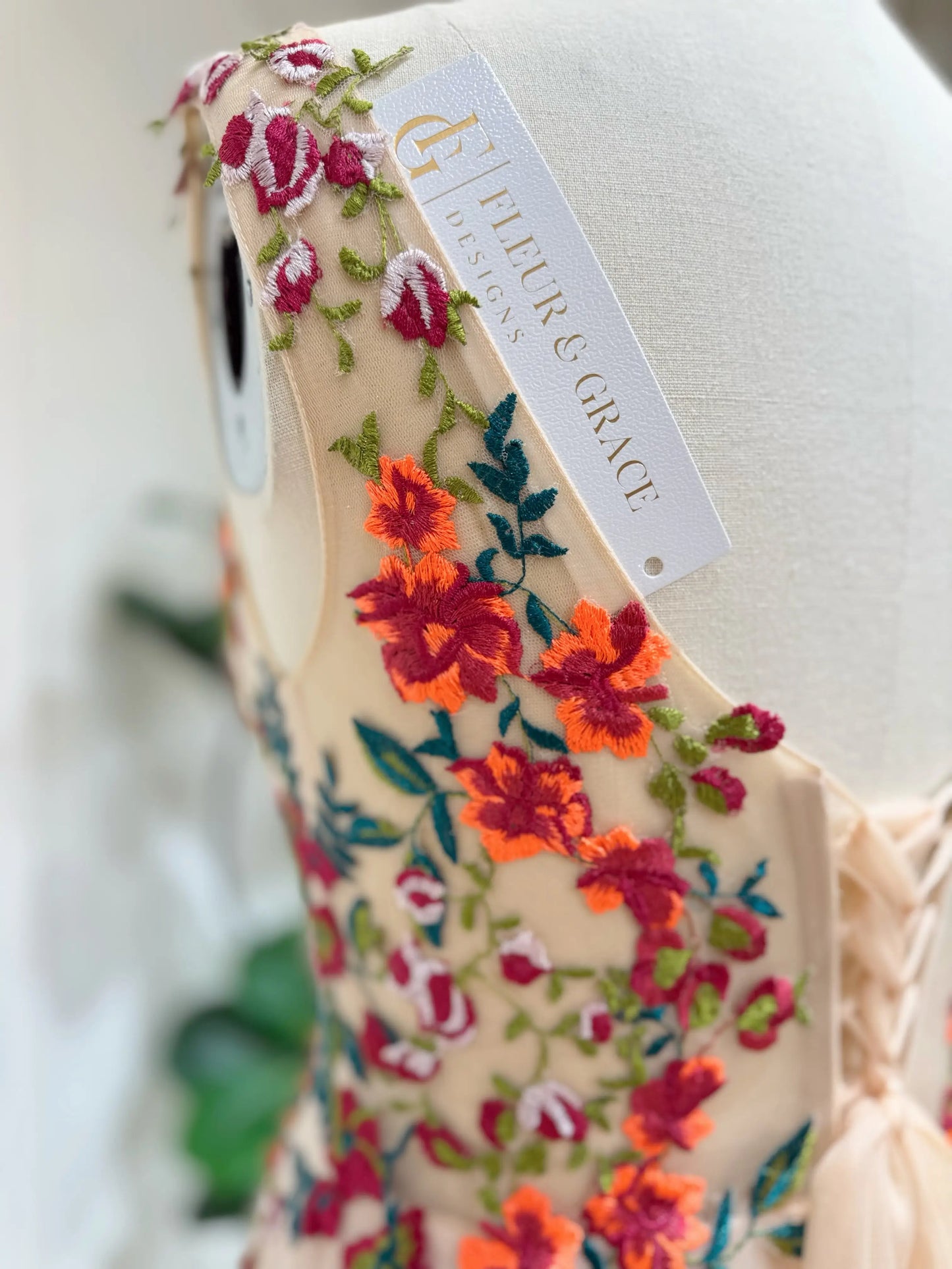 Bride mannequin wearing the Monica Gown by Fleur&Grace Designs — colorful wedding dress with orange floral appliqués, meadow green embroidery on champagne tulle, V-neck, sleeveless bodice, lace-up back, and pockets.