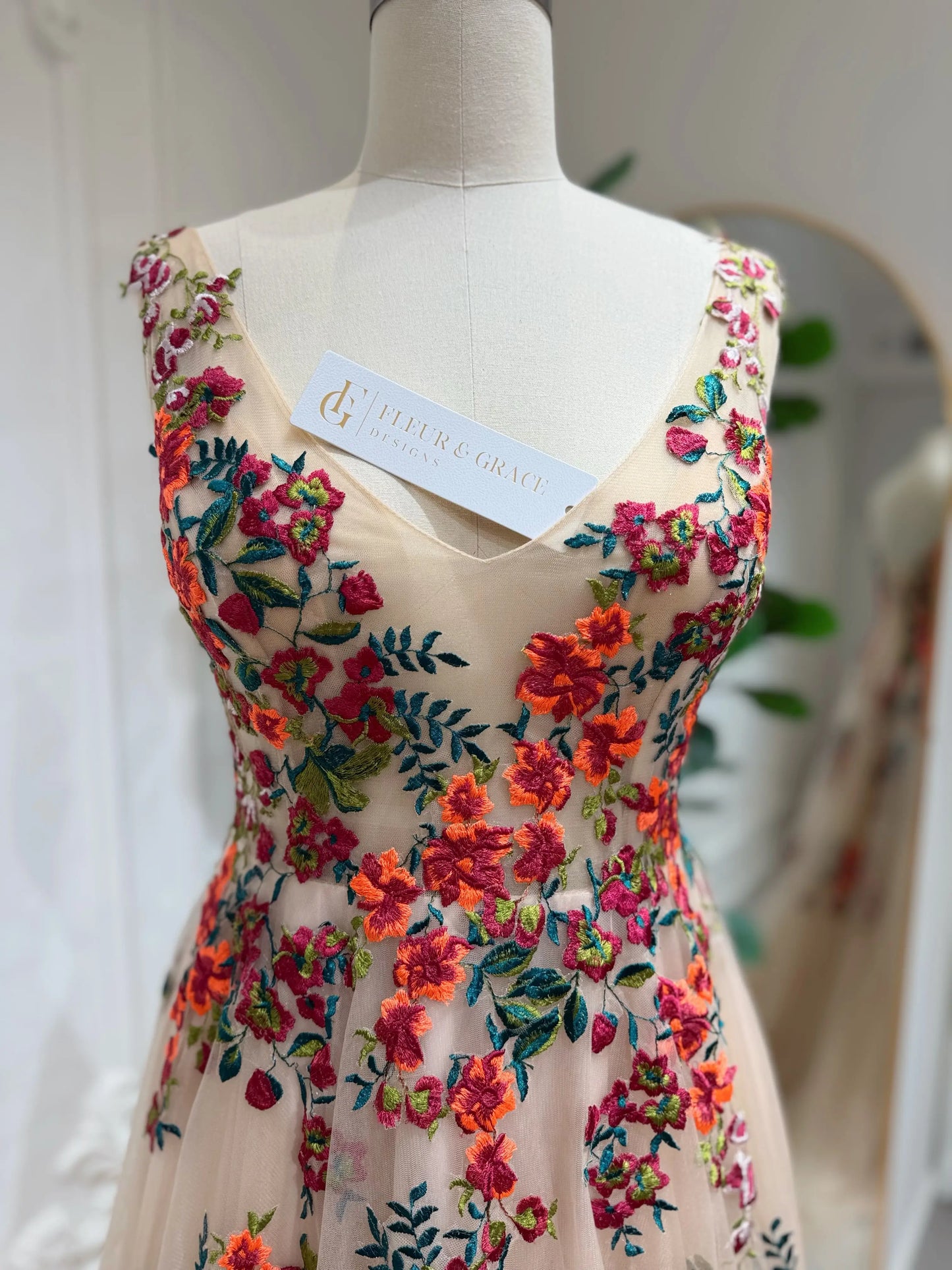Bride mannequin wearing the Monica Gown by Fleur&Grace Designs — colorful wedding dress with orange floral appliqués, meadow green embroidery on champagne tulle, V-neck, sleeveless bodice, lace-up back, and pockets.