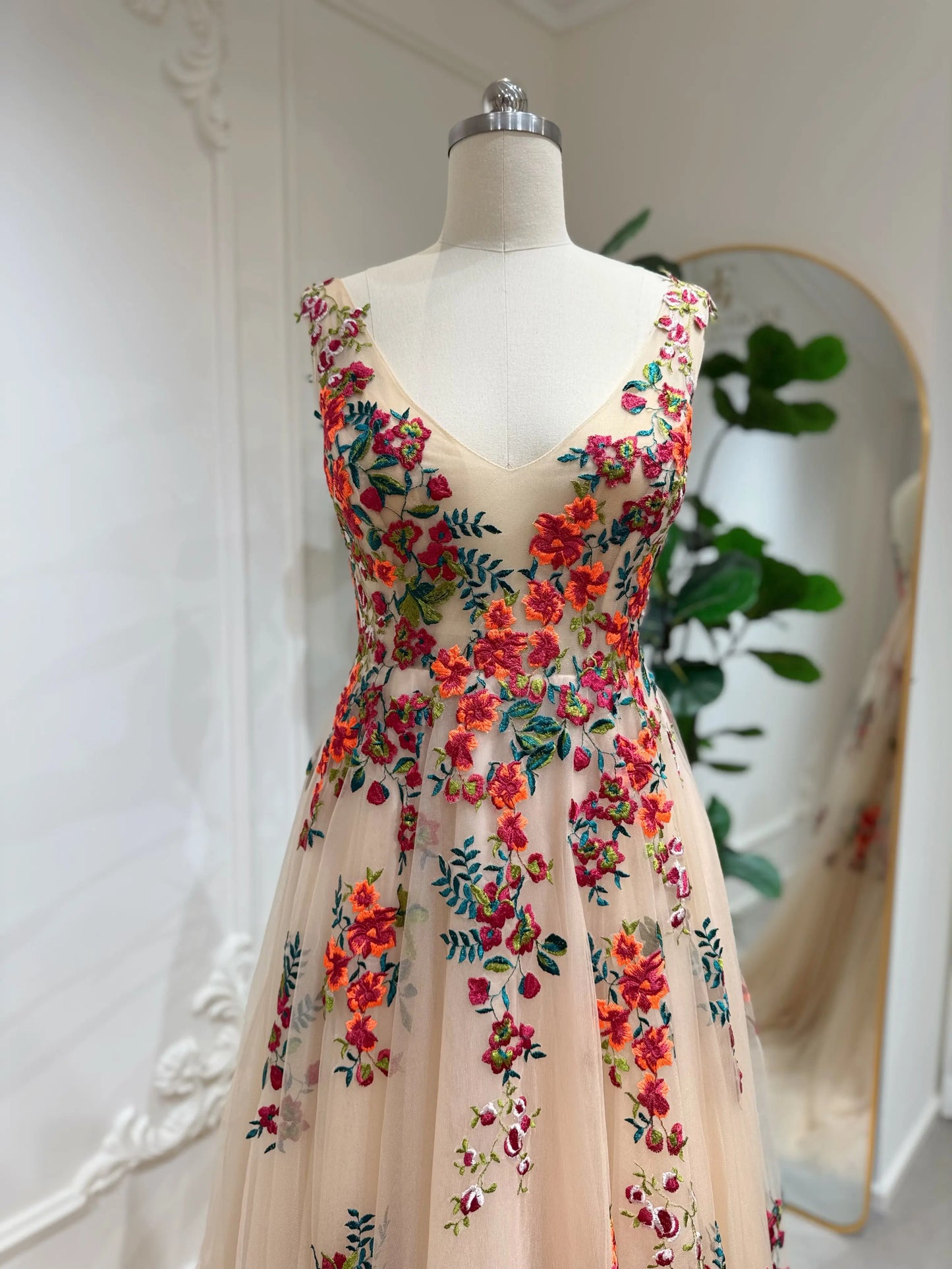 Bride mannequin wearing the Monica Gown by Fleur&Grace Designs — colorful wedding dress with orange floral appliqués, meadow green embroidery on champagne tulle, V-neck, sleeveless bodice, lace-up back, and pockets.