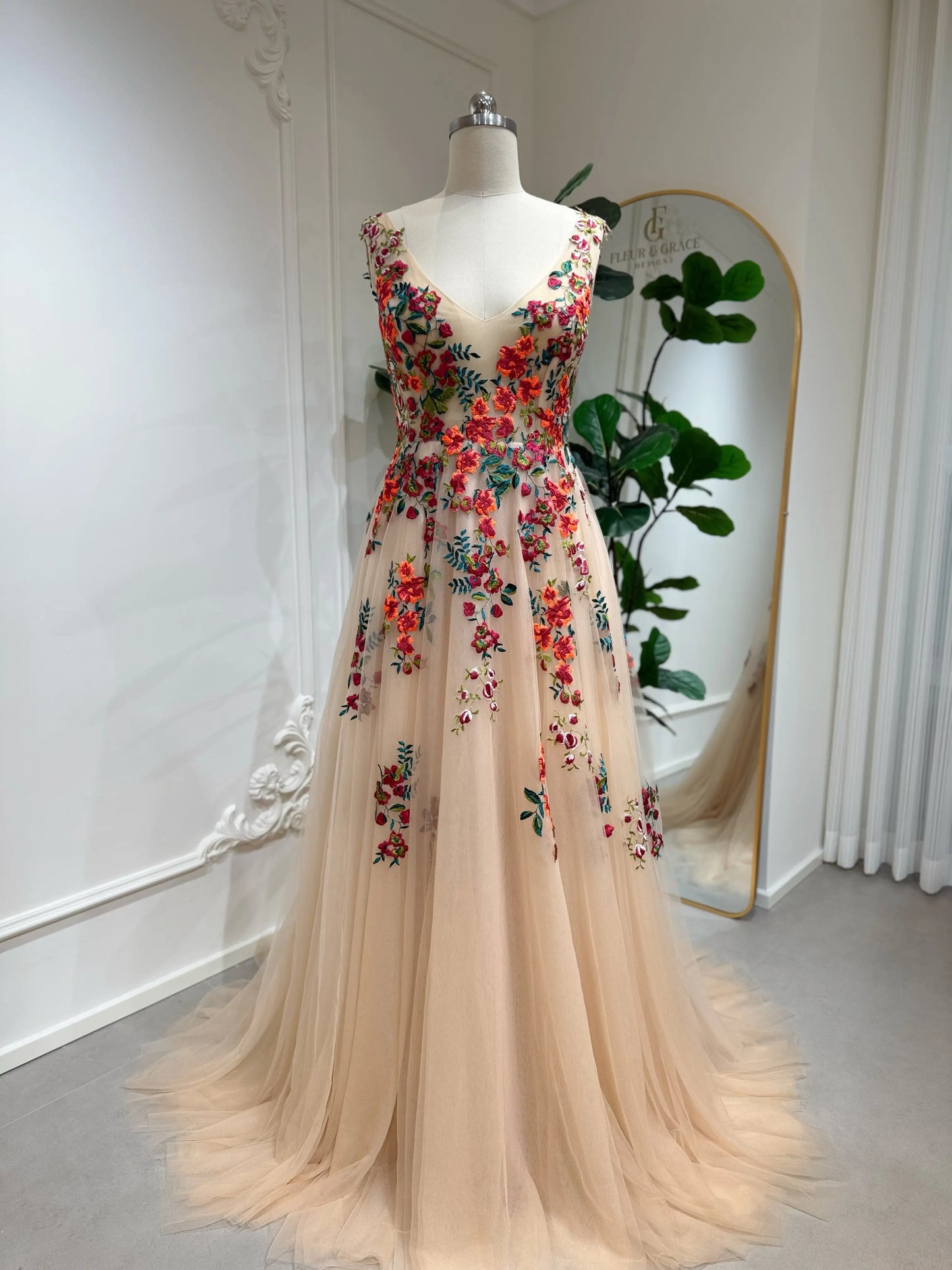 Bride mannequin wearing the Monica Gown by Fleur&Grace Designs — colorful wedding dress with orange floral appliqués, meadow green embroidery on champagne tulle, V-neck, sleeveless bodice, lace-up back, and pockets.