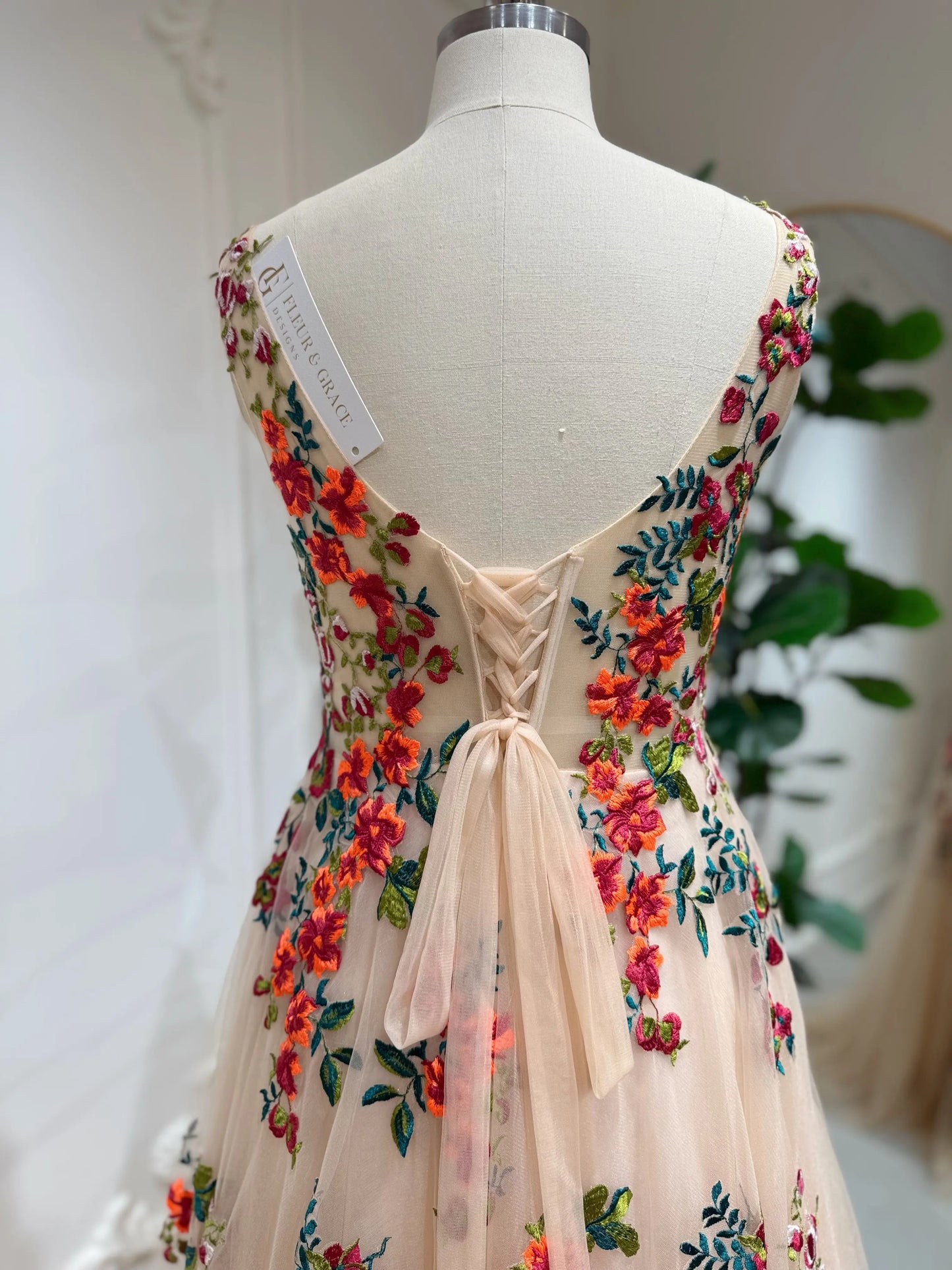 Bride mannequin wearing the Monica Gown by Fleur&Grace Designs — colorful wedding dress with orange floral appliqués, meadow green embroidery on champagne tulle, V-neck, sleeveless bodice, lace-up back, and pockets.