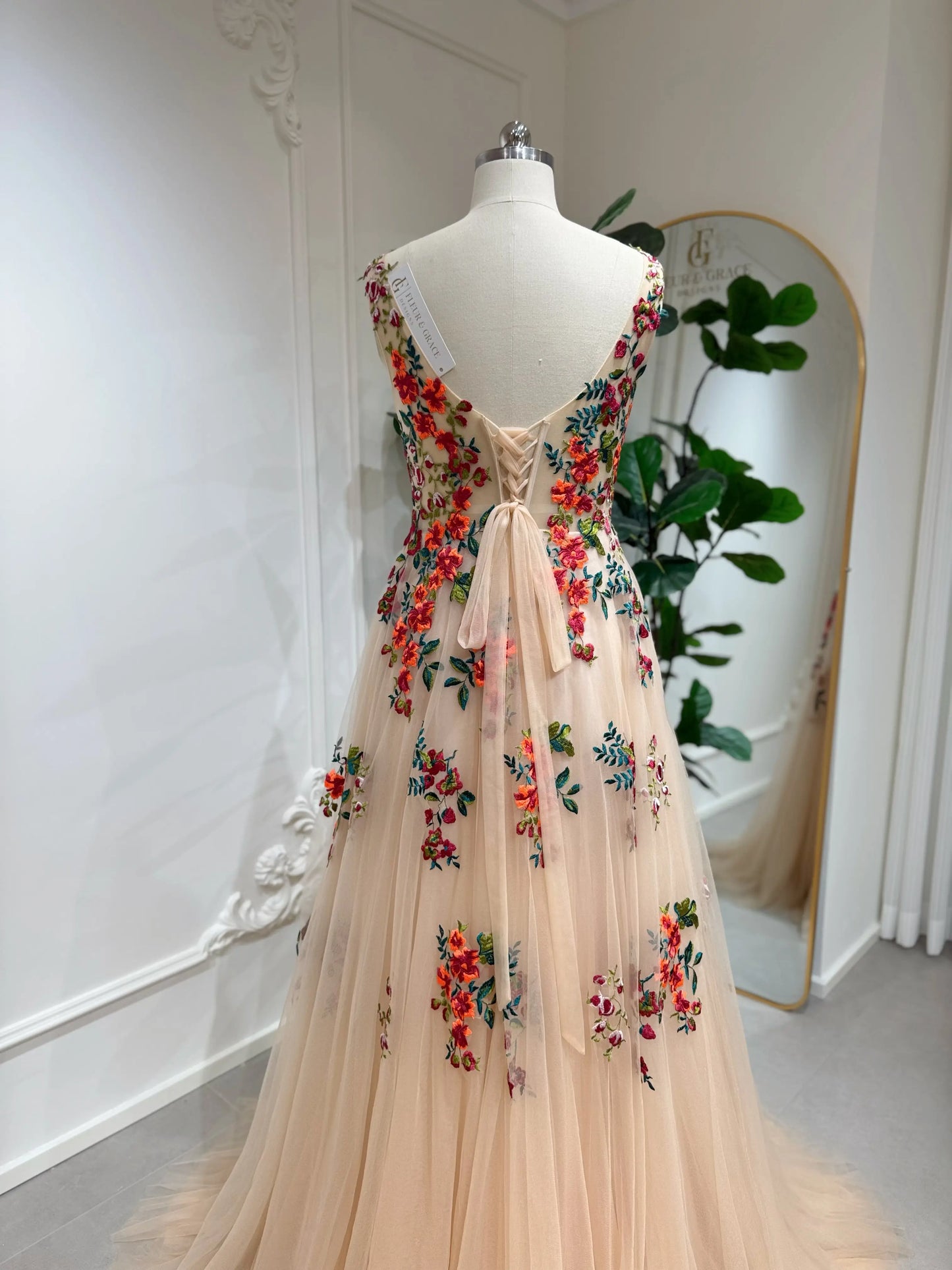 Bride mannequin wearing the Monica Gown by Fleur&Grace Designs — colorful wedding dress with orange floral appliqués, meadow green embroidery on champagne tulle, V-neck, sleeveless bodice, lace-up back, and pockets.