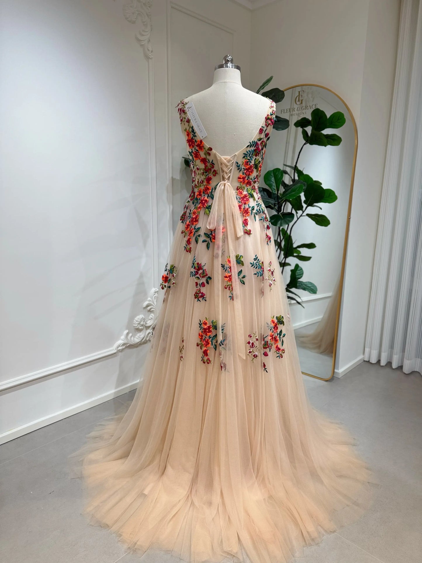 Bride mannequin wearing the Monica Gown by Fleur&Grace Designs — colorful wedding dress with orange floral appliqués, meadow green embroidery on champagne tulle, V-neck, sleeveless bodice, lace-up back, and pockets.