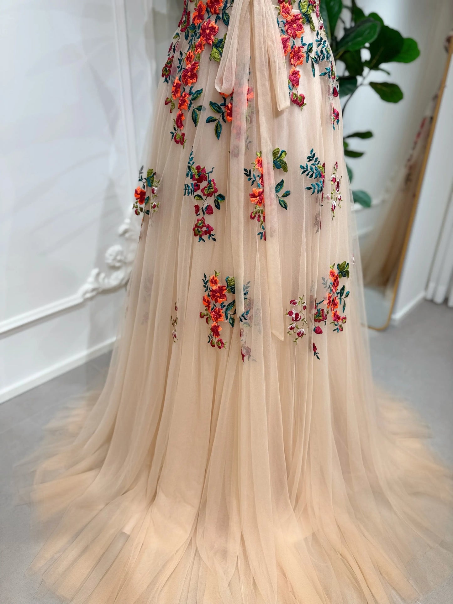 Bride mannequin wearing the Monica Gown by Fleur&Grace Designs — colorful wedding dress with orange floral appliqués, meadow green embroidery on champagne tulle, V-neck, sleeveless bodice, lace-up back, and pockets.