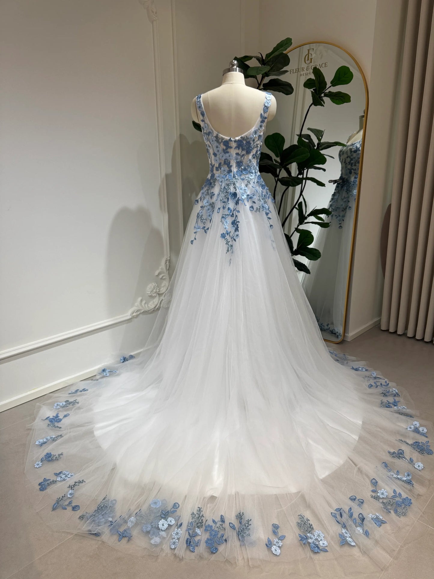 The Iris Gown by Fleur&Grace Designs is a graceful and romantic wedding dress adorned with elegant dusty blue floral embroidery. Crafted in soft tulle with a structured silhouette, this gown features a timeless square neckline, a comfortable built-in bodice, and pockets for modern functionality.