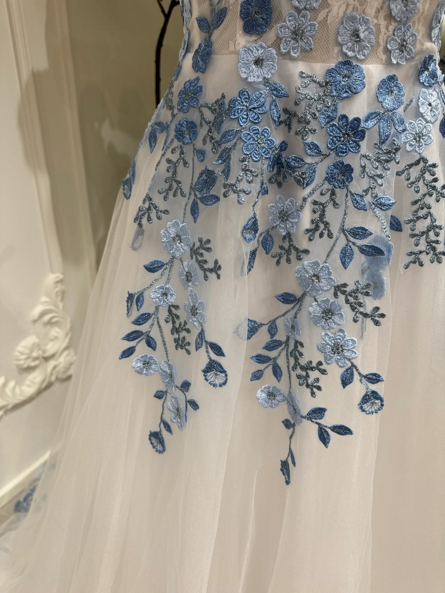 The Iris Gown by Fleur&Grace Designs is a graceful and romantic wedding dress adorned with elegant dusty blue floral embroidery. Crafted in soft tulle with a structured silhouette, this gown features a timeless square neckline, a comfortable built-in bodice, and pockets for modern functionality.