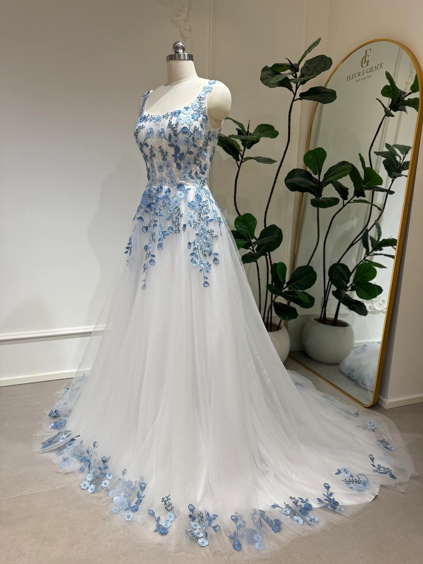 The Iris Gown by Fleur&Grace Designs is a graceful and romantic wedding dress adorned with elegant dusty blue floral embroidery. Crafted in soft tulle with a structured silhouette, this gown features a timeless square neckline, a comfortable built-in bodice, and pockets for modern functionality.