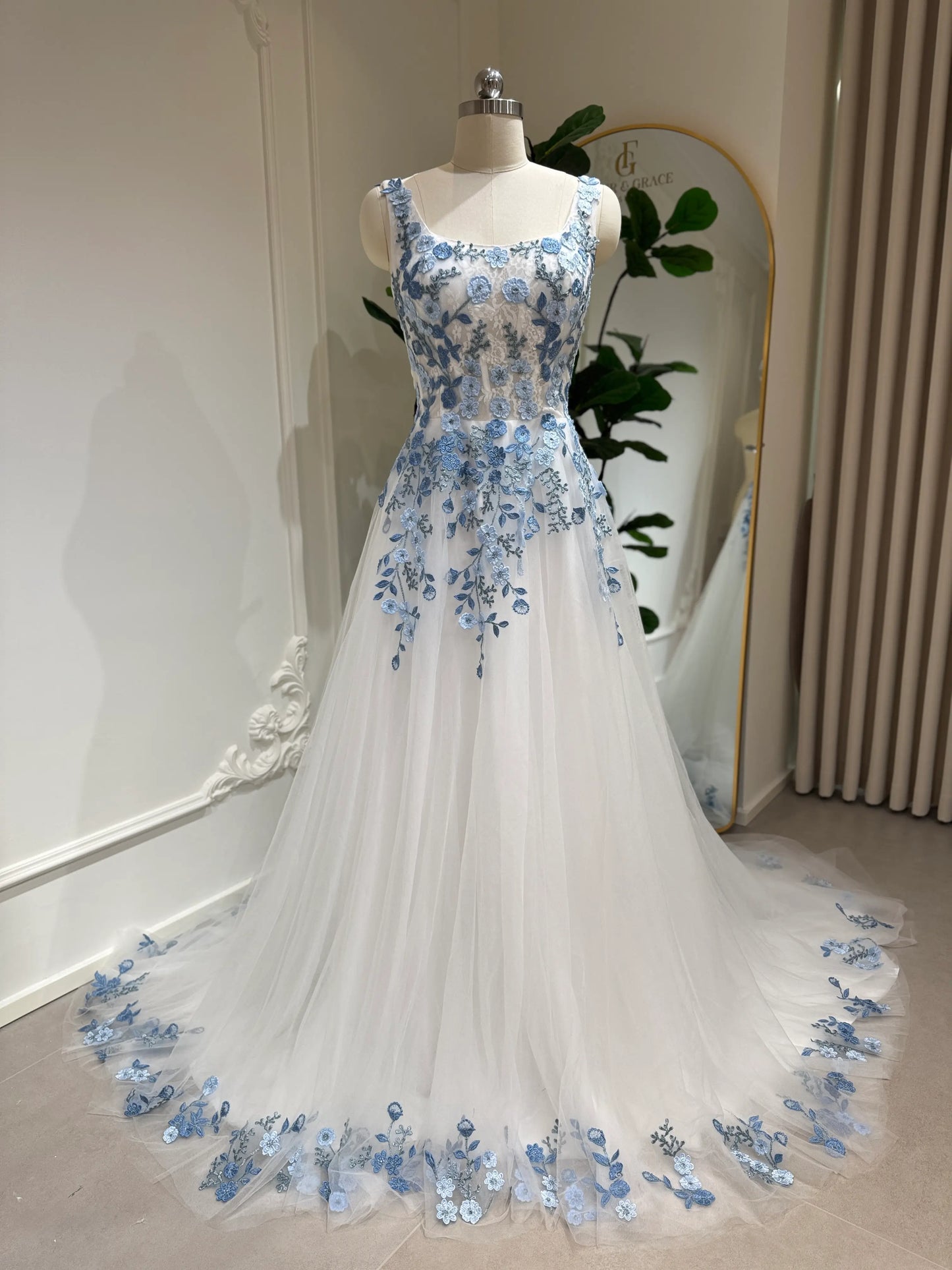 The Iris Gown by Fleur&Grace Designs is a graceful and romantic wedding dress adorned with elegant dusty blue floral embroidery. Crafted in soft tulle with a structured silhouette, this gown features a timeless square neckline, a comfortable built-in bodice, and pockets for modern functionality.
