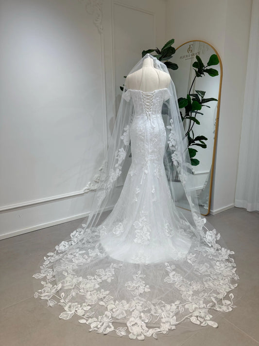 Bridal mannequin wearing the Lena Gown FG030 by Fleur&Grace Designs—a mermaid lace wedding dress with off-the-shoulder sleeves, sheer boned bodice, corset back, basque waistline, asymmetric hemline, and chapel-length train.