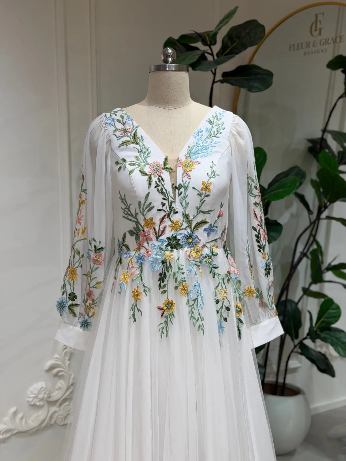 White wedding dress with colorful floral embroidery, V-neckline, and sheer long sleeves on a mannequin.