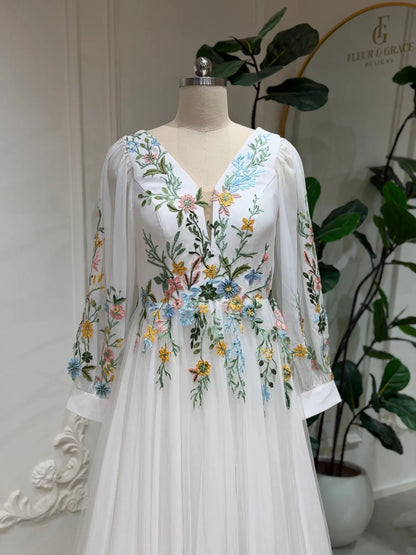 White wedding dress with colorful floral embroidery, V-neckline, and sheer long sleeves on a mannequin.