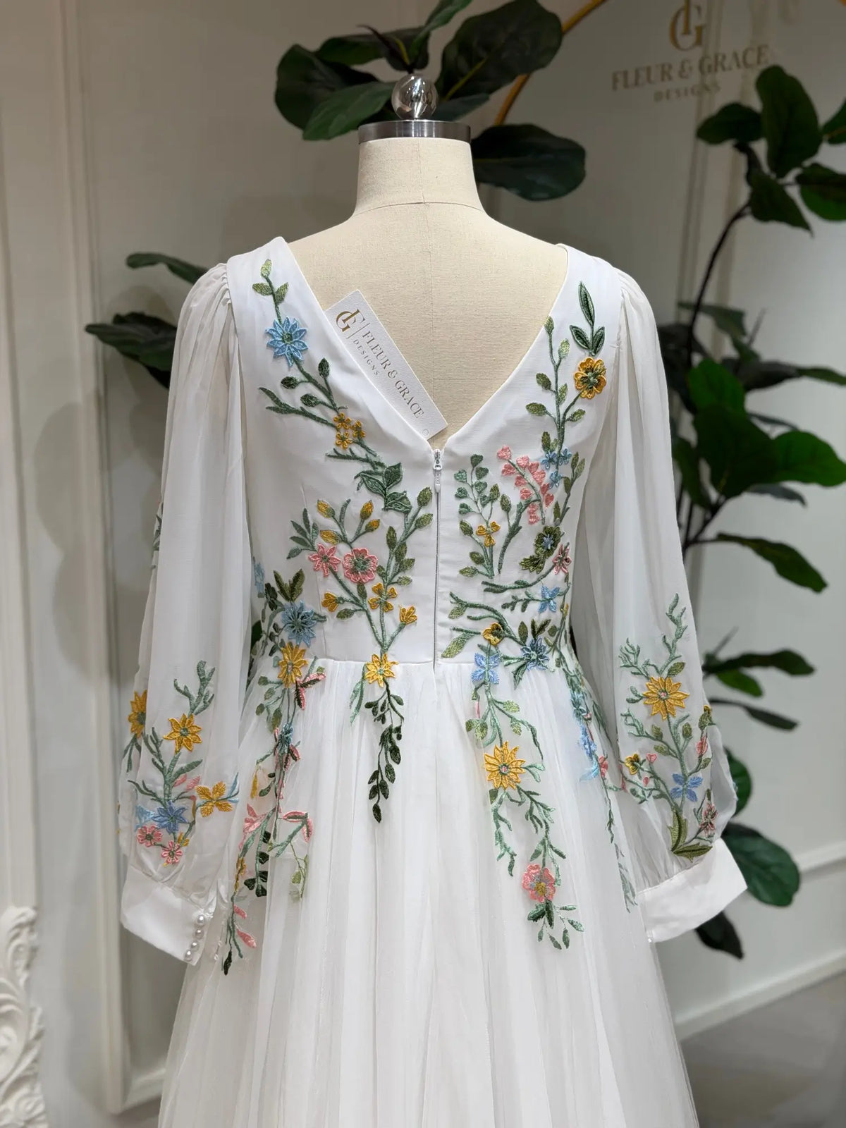 White wedding dress with colorful floral embroidery, V-back, and long sleeves on a mannequin.