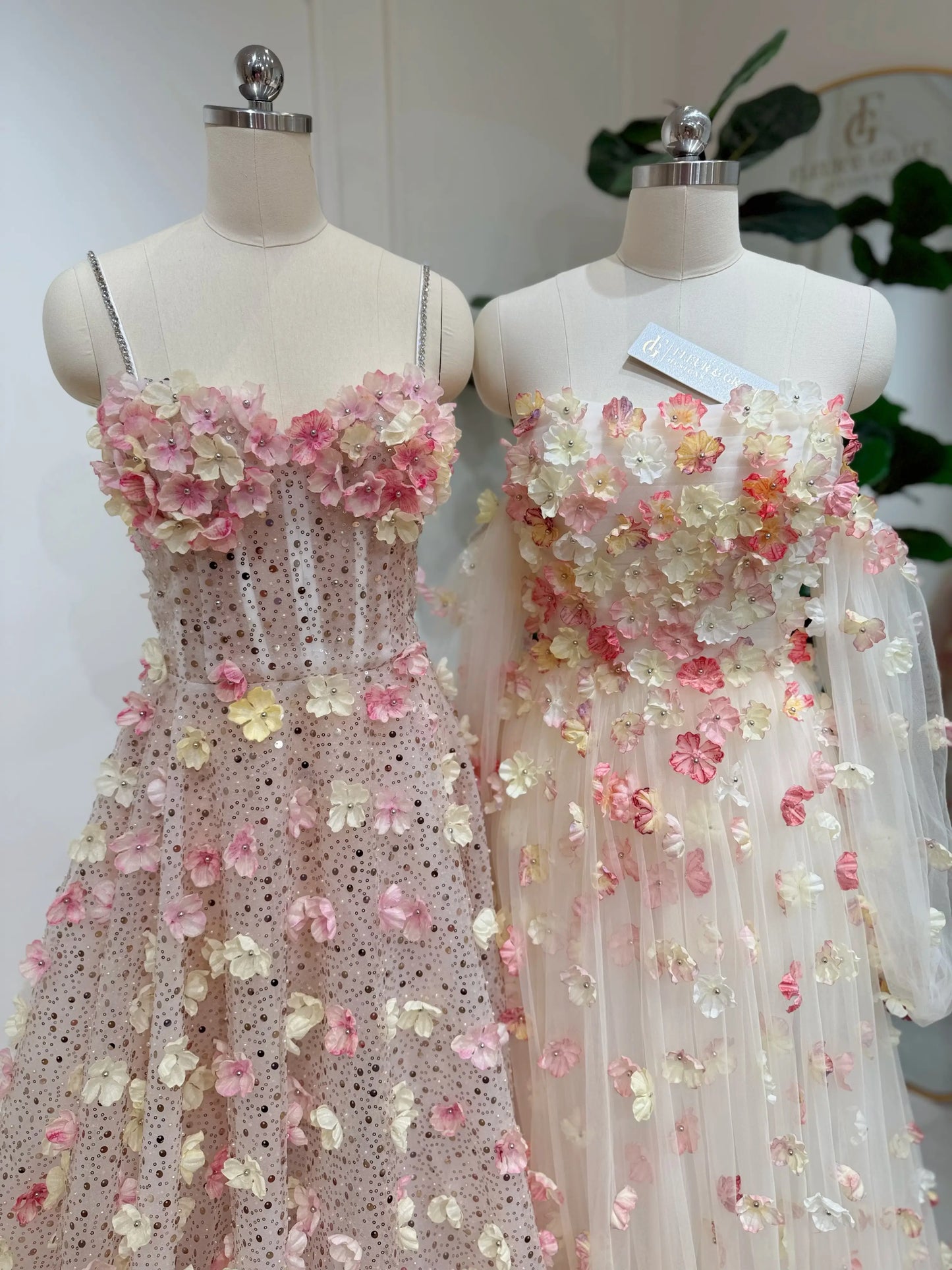 Two mannequins wearing pink and yellow 3D floral lace gowns with sweetheart necklines and shimmering beaded details.