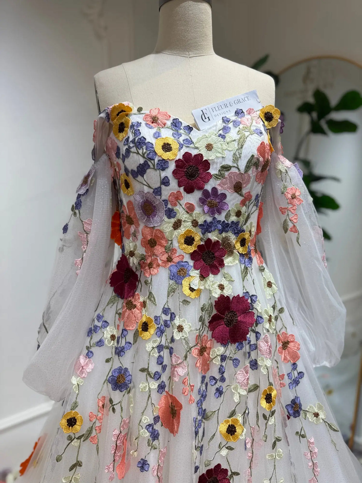 Off-the-shoulder wedding dress with colorful 3D floral embroidery, sheer bishop sleeves, and sparkle tulle fabric.