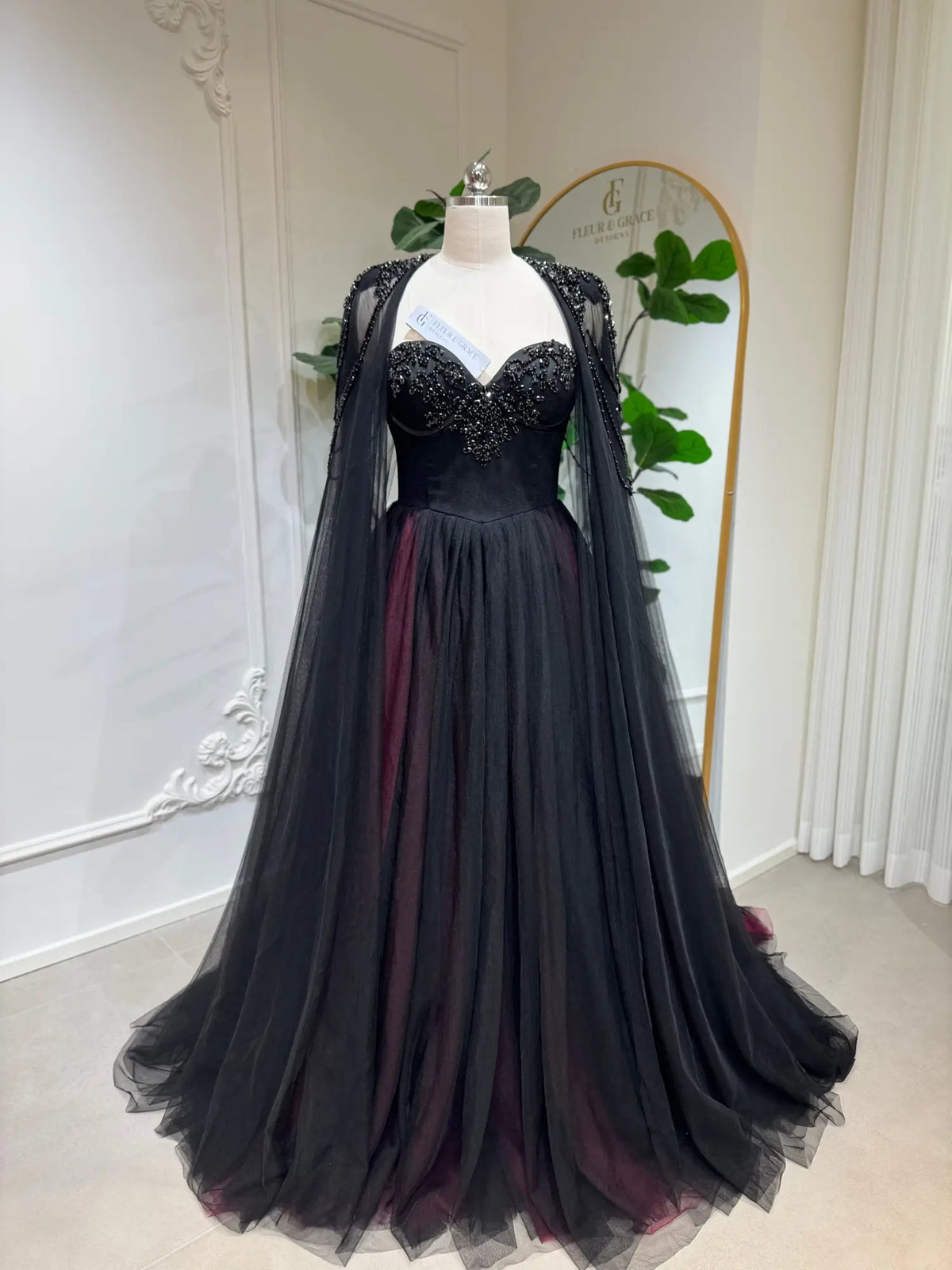 Black and burgundy A-line wedding dress with a beaded corset bodice and detachable illusion cape on a mannequin.