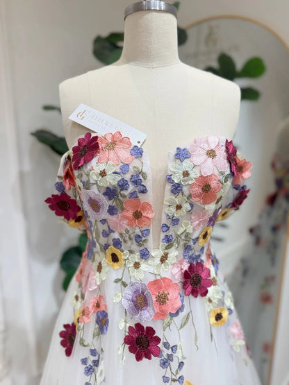 Off-the-shoulder wedding gown in off-white tulle with vibrant multicolored floral embroidery and a plunging V-neck bodice.