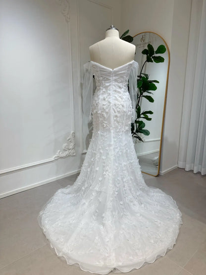 White mermaid wedding dress with 3D floral appliqués, off-the-shoulder illusion sleeves, and a flowing train.