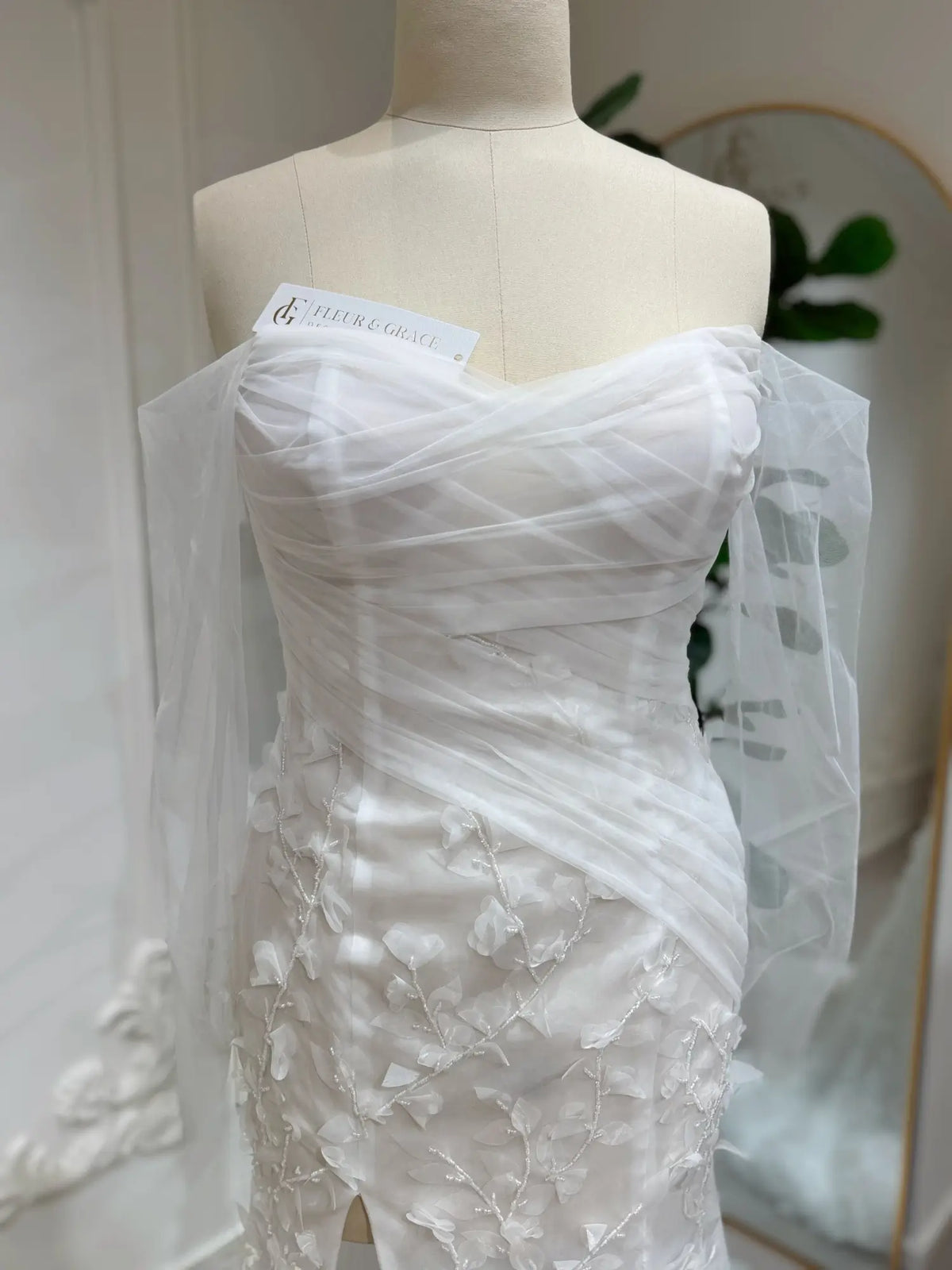 White mermaid wedding dress with 3D floral appliqués, beaded details, and draped off-the-shoulder illusion sleeves.