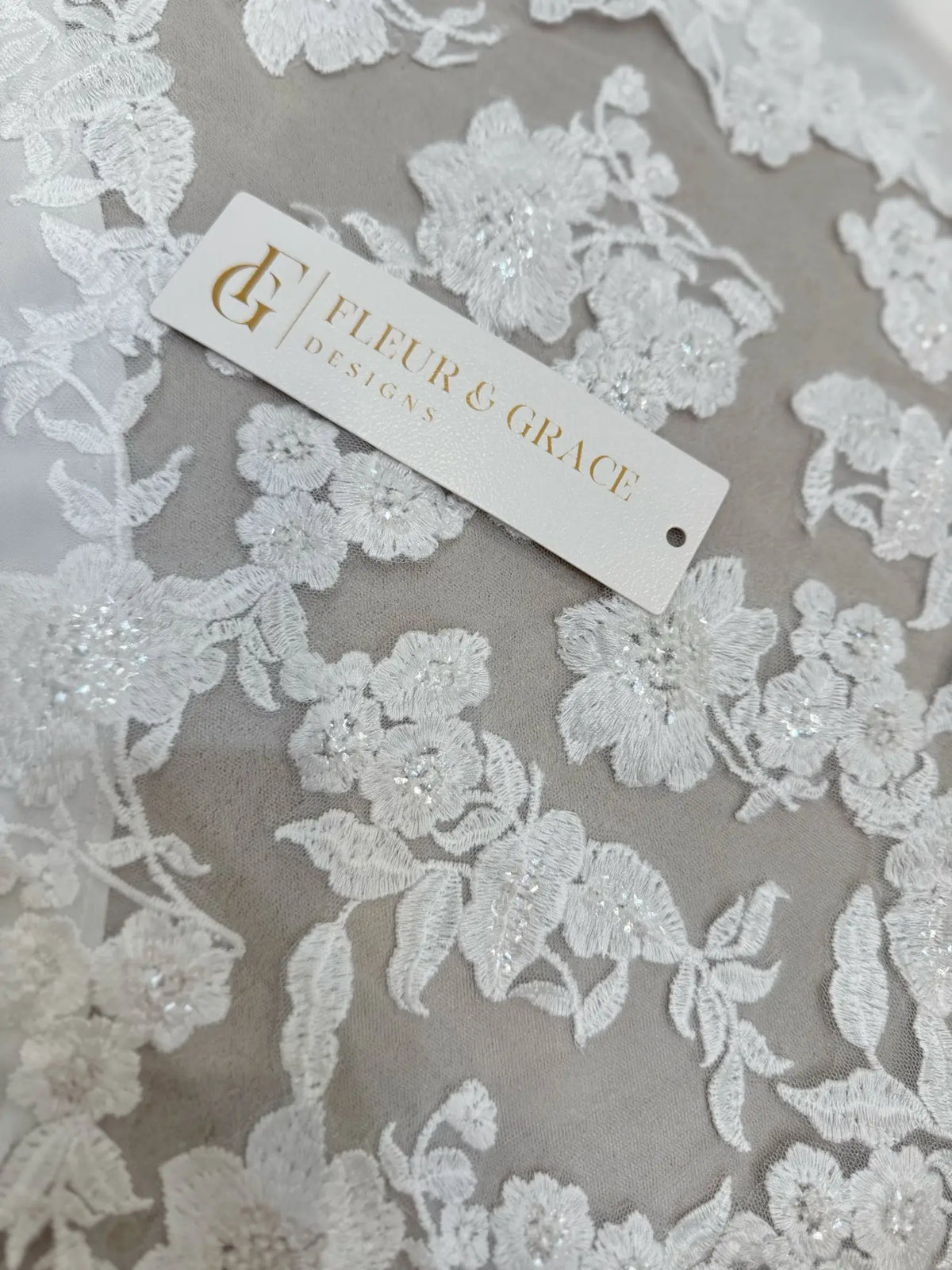 White floral lace appliqué with delicate beading on soft chiffon fabric, featuring a Fleur & Grace brand tag.