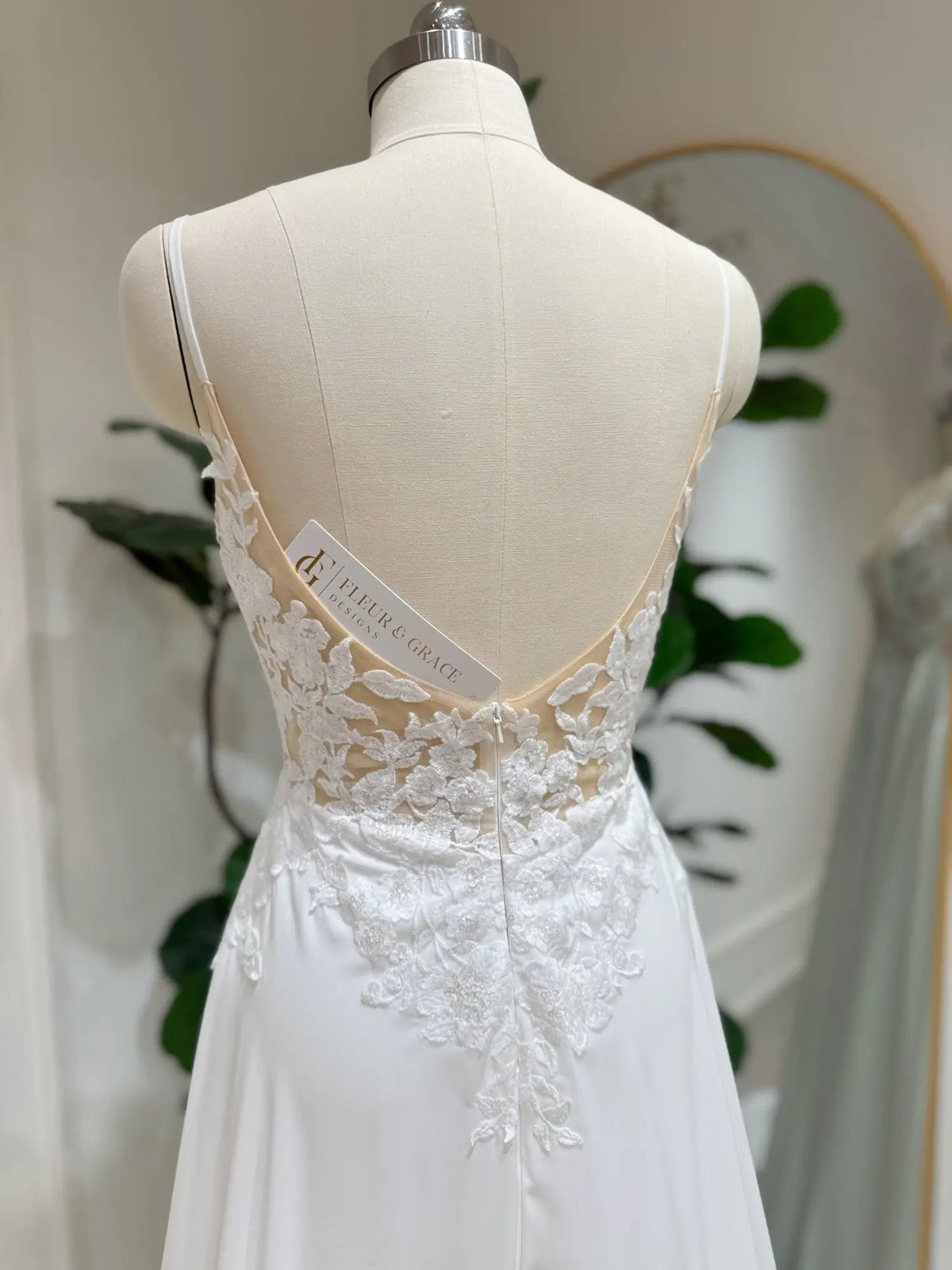 White chiffon wedding dress on a mannequin featuring a low open back, delicate floral lace appliqué, and thin straps.