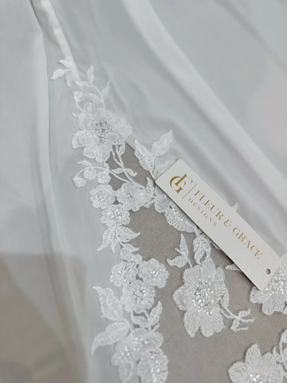 White chiffon wedding dress featuring intricate floral lace appliqué and a Fleur & Grace brand tag.