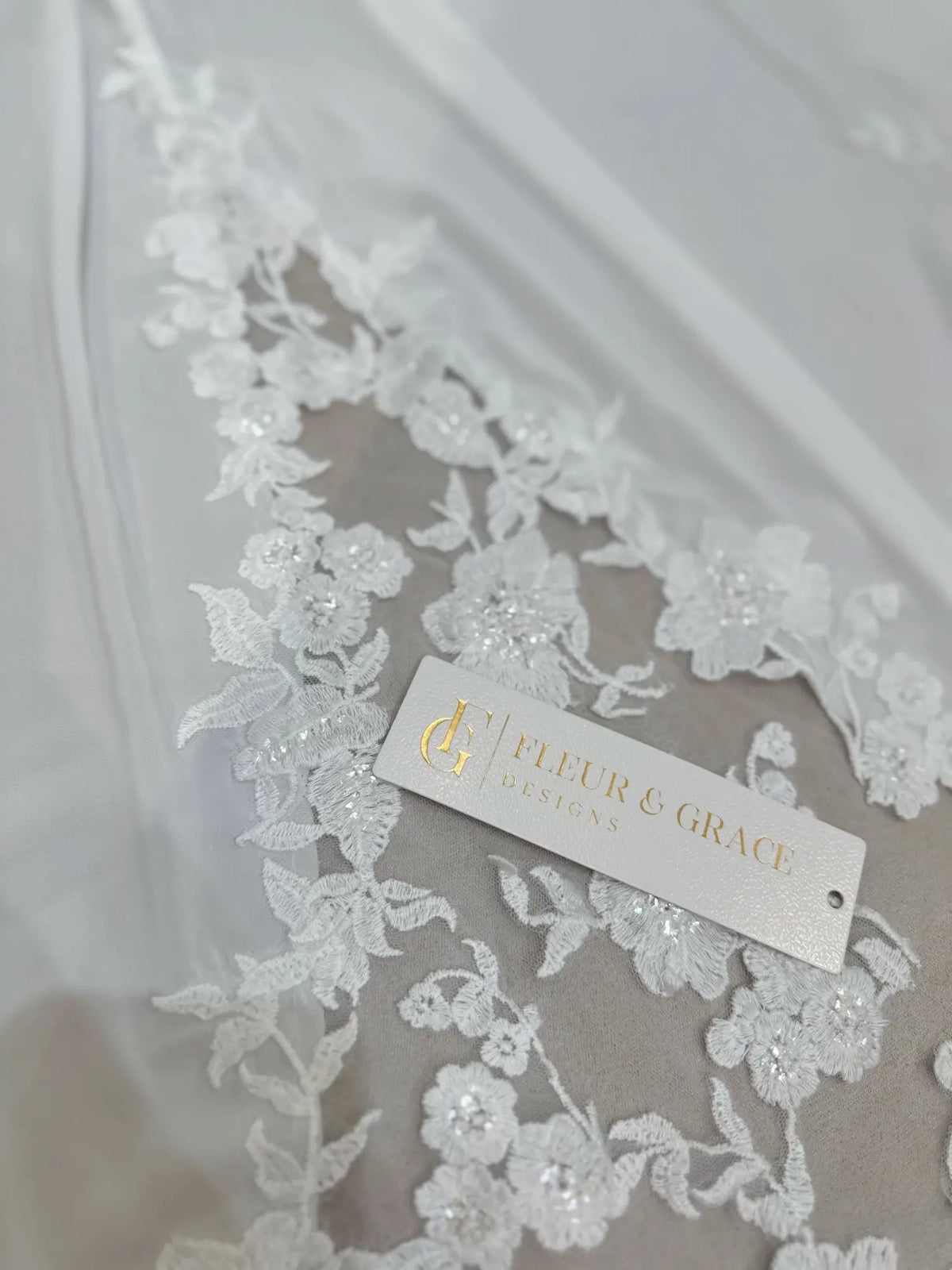 White chiffon wedding dress fabric featuring delicate floral lace appliqué with subtle beadwork and a branded tag.