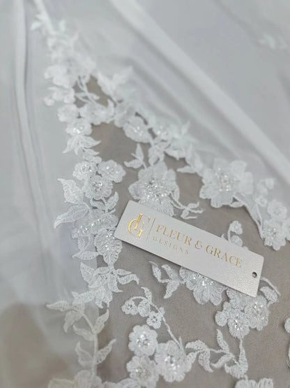 White chiffon wedding dress fabric featuring delicate floral lace appliqué with subtle beadwork and a branded tag.