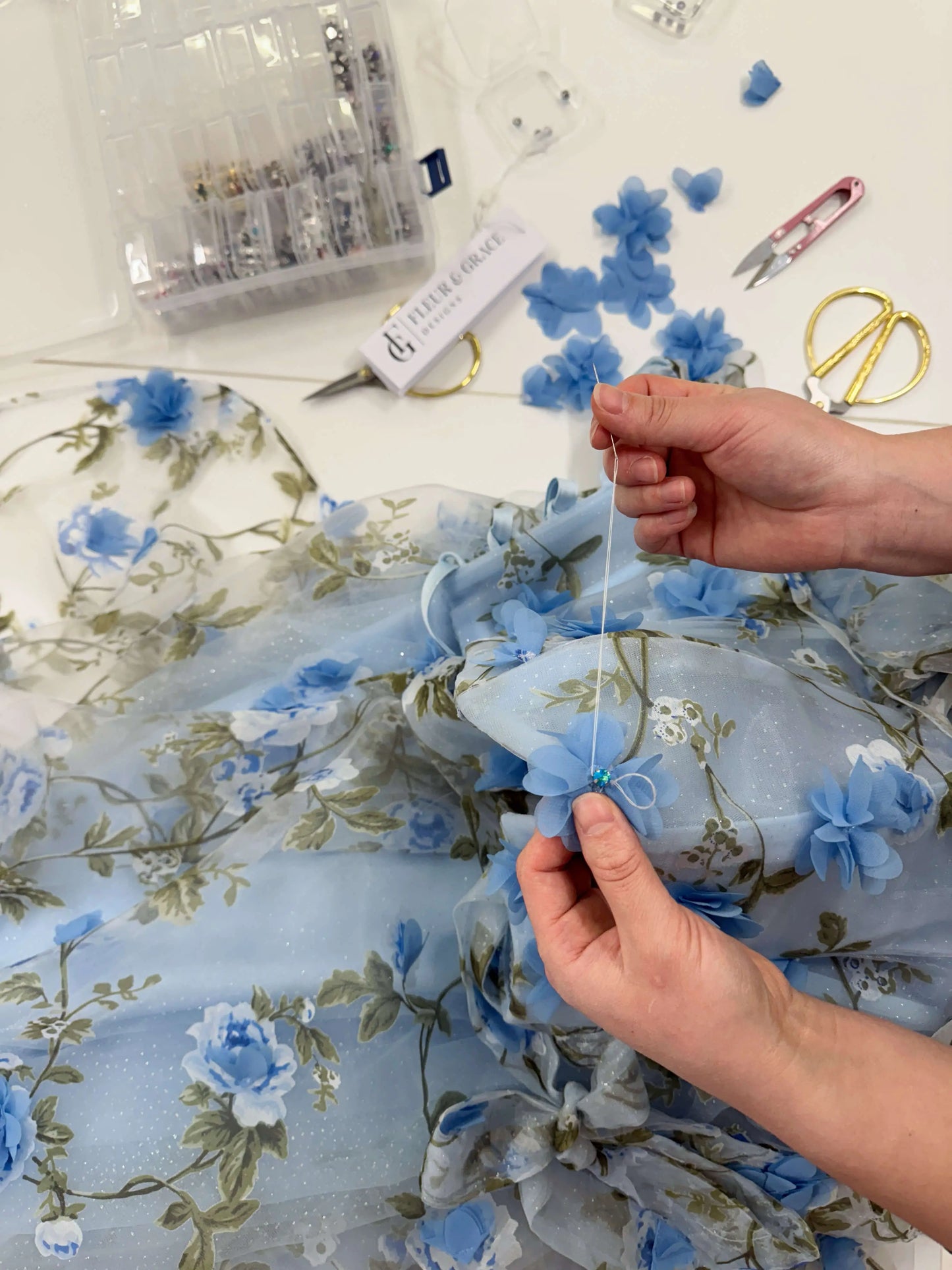 Hands sewing 3D blue floral organza lace onto a sparkly mesh gown with green leaf prints.
