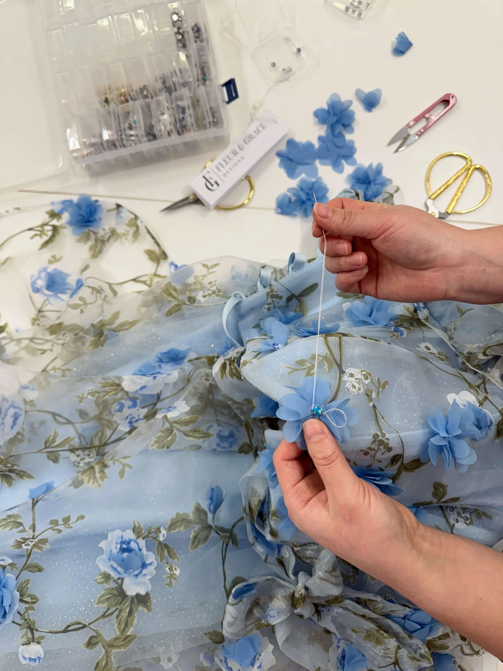 Hands sewing 3D blue floral organza lace onto a sparkly mesh gown with green leaf prints.