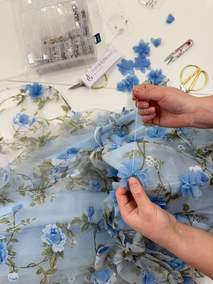Hands sewing 3D blue floral organza lace onto a sparkly mesh gown with green leaf prints.