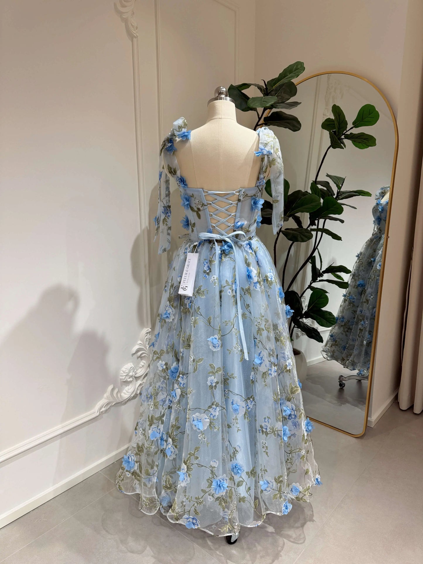Blue midi gown with 3D floral organza, green leaf prints, sweetheart neckline, and lace-up back on a mannequin.