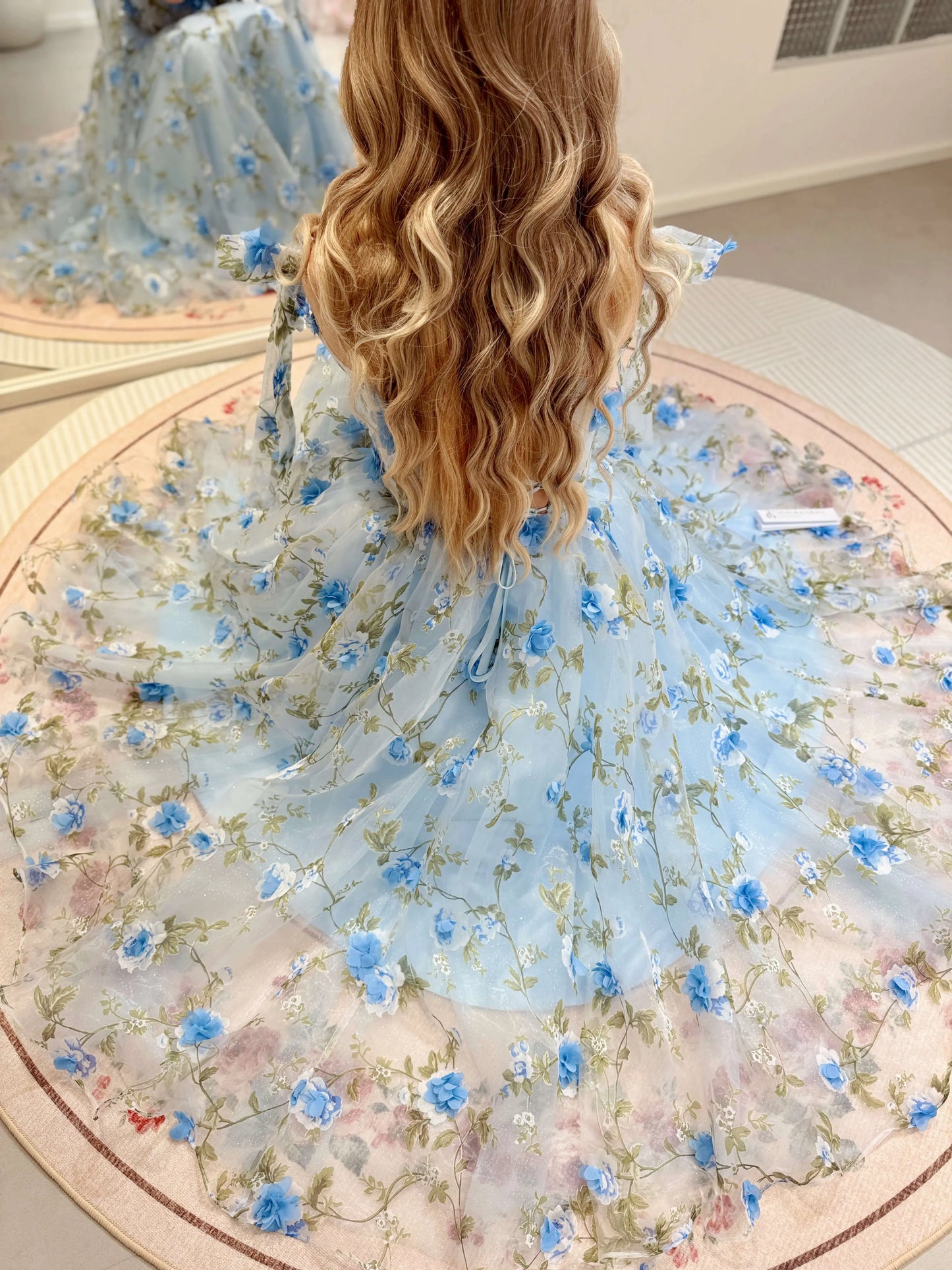 Blue midi gown with 3D floral organza, green leaf prints, and sparkly mesh, featuring an adjustable lace-up back design.