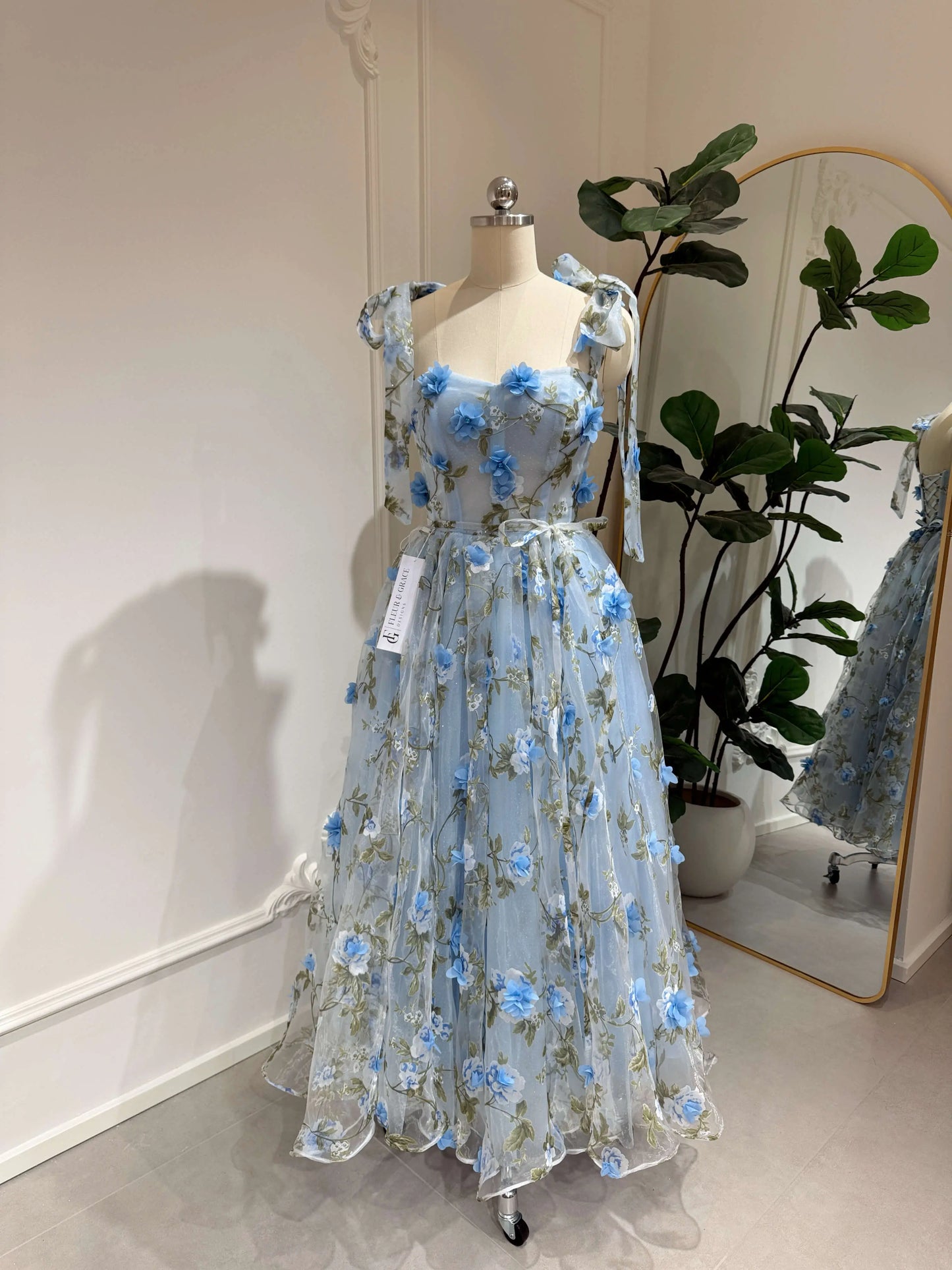 Blue midi gown with 3D floral organza, green leaf prints, sweetheart neckline, and tie-shoulder straps on a mannequin.