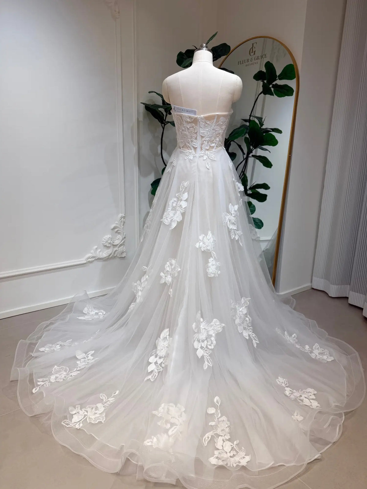 Strapless white A-line wedding dress with 3D floral lace appliqués and a flowing tulle chapel train on a mannequin.