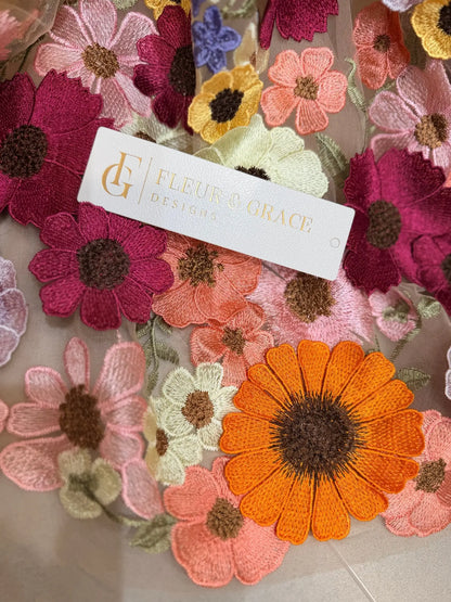 Close-up of 3D floral embroidery in vibrant orange, pink, and yellow on sheer tulle fabric with a Fleur & Grace brand tag.