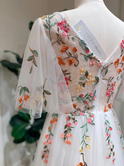 White tulle wedding dress with colorful floral embroidery, V-neckline, and airy flutter sleeves on a mannequin.