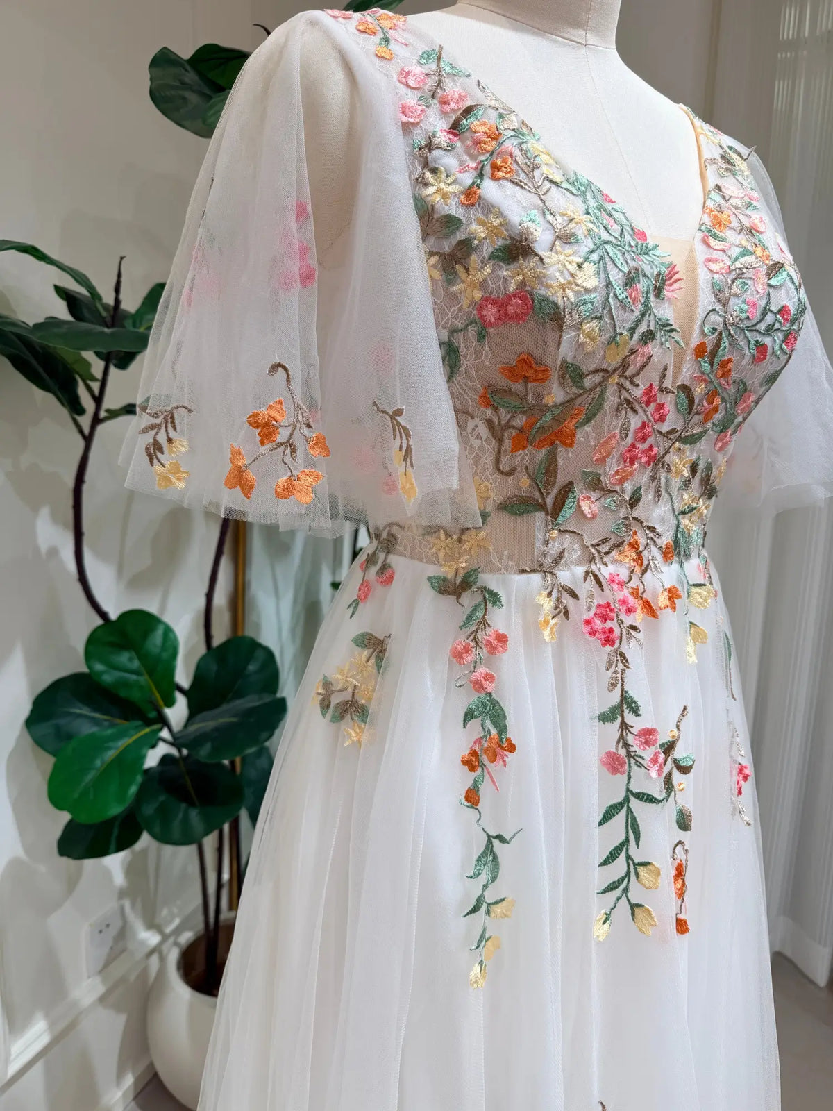 White tulle wedding dress with colorful floral embroidery, V-neckline, and airy flutter sleeves on a mannequin.