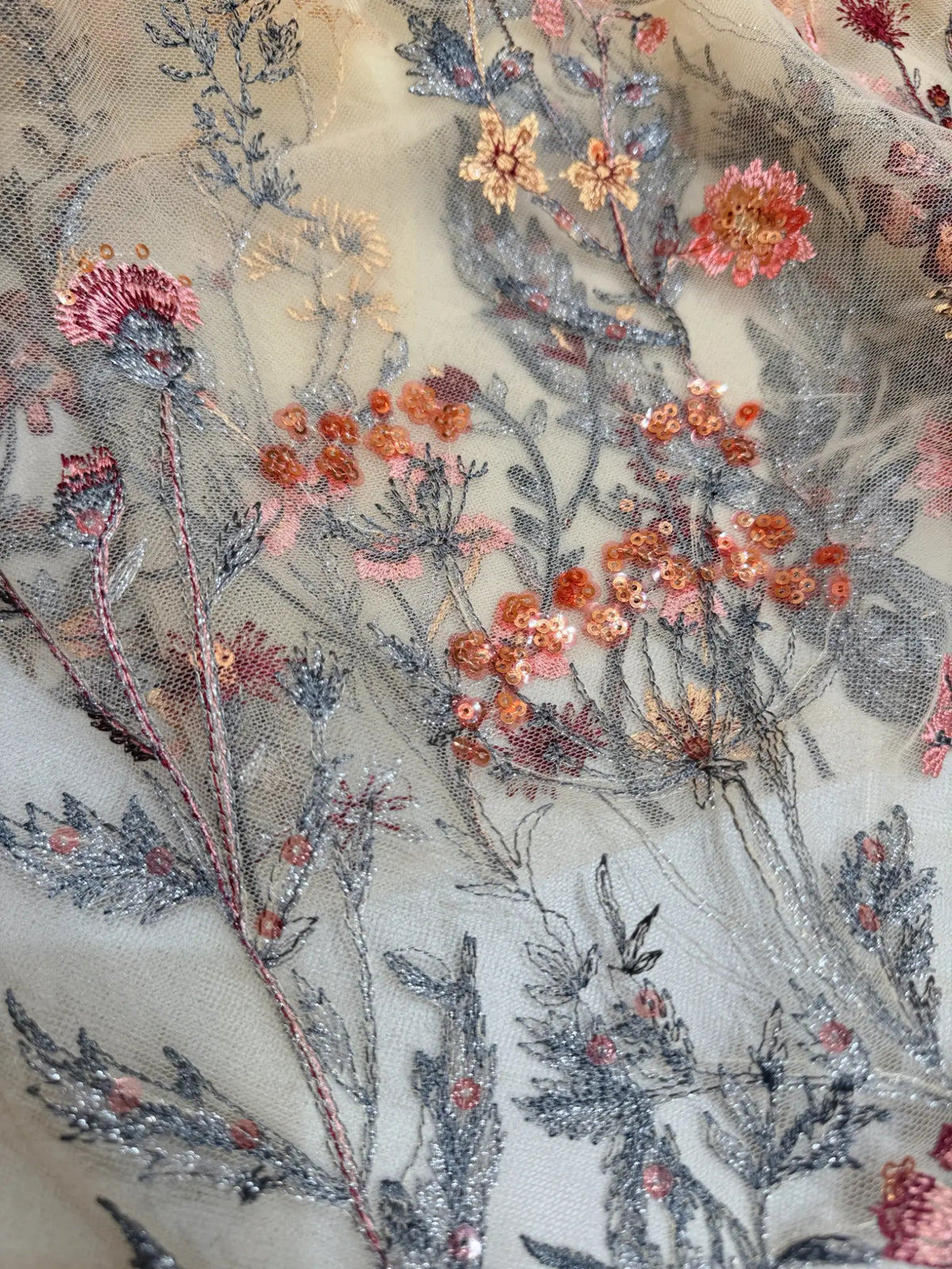 Sheer tulle fabric featuring intricate wildflower meadow embroidery with pink, orange, and metallic silver floral details.