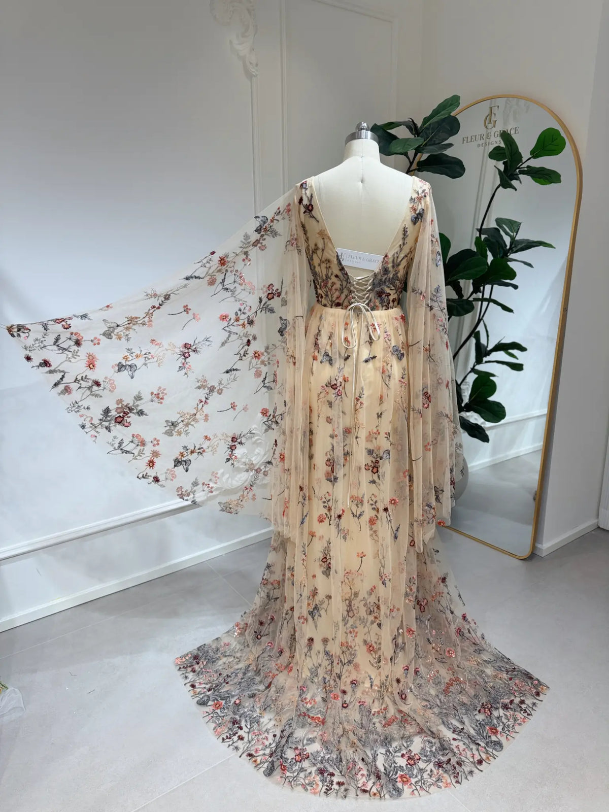 Backless botanical wedding dress with wildflower lace embroidery, flowing cape sleeves, and a lace-up corset back.