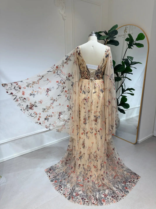 Backless botanical wedding dress with wildflower lace embroidery, flowing cape sleeves, and a lace-up corset back.