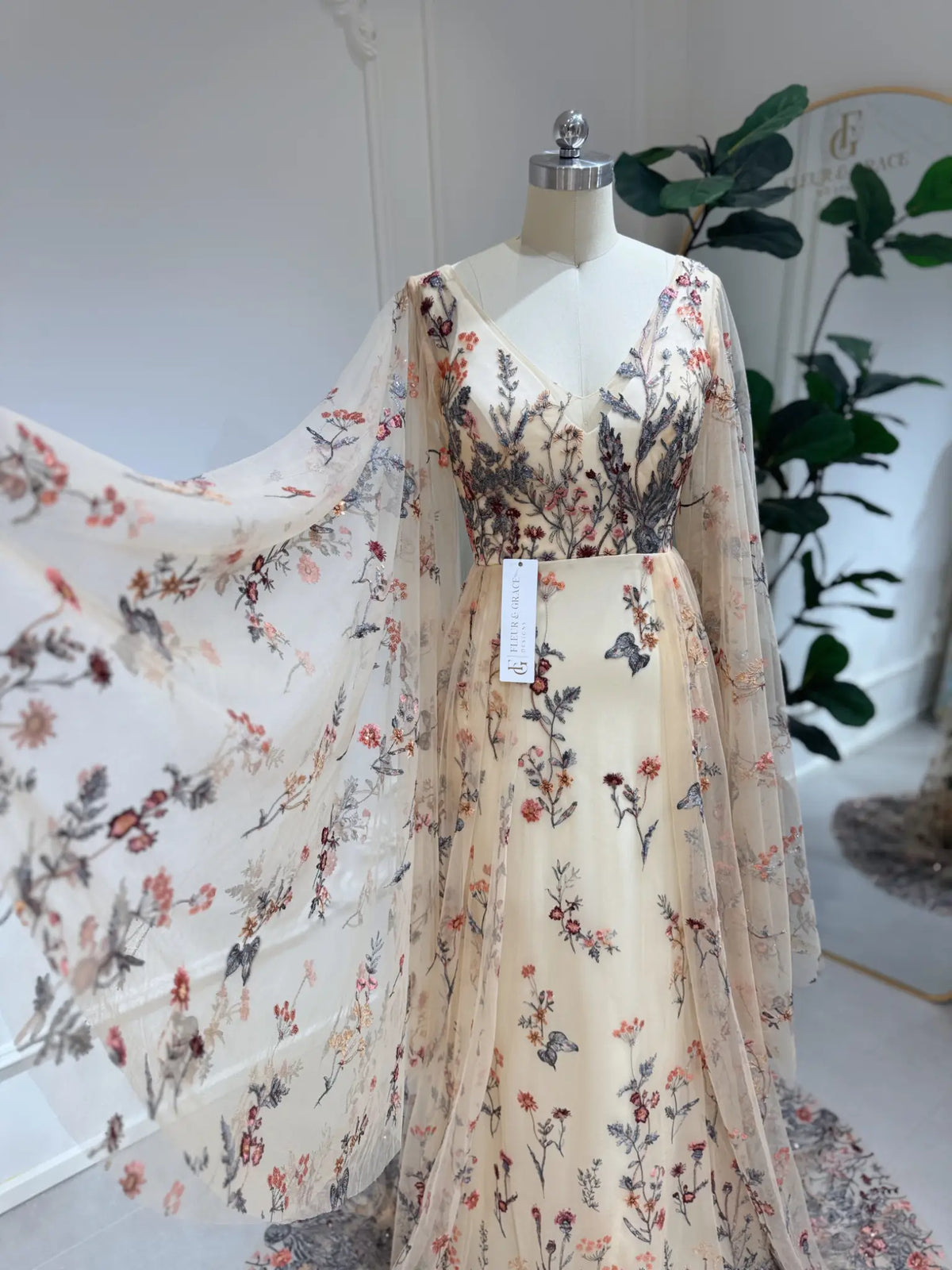 Elva botanical wedding dress with sheer tulle, wildflower embroidery, V-neckline, and dramatic cape sleeves on a mannequin.
