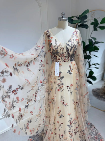 Elva botanical wedding dress with sheer tulle, wildflower embroidery, V-neckline, and dramatic cape sleeves on a mannequin.