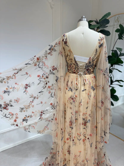 Backless botanical wedding dress with wildflower embroidery, sheer cape sleeves, and a lace-up corset back on a mannequin.