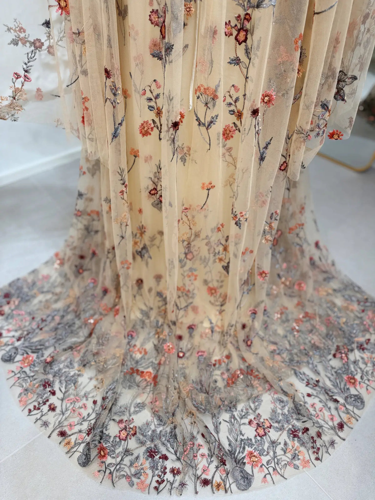 Beige tulle wedding dress with intricate wildflower meadow embroidery and flowing cape sleeves.