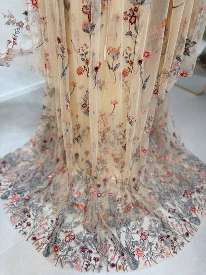 Beige tulle wedding dress with intricate wildflower meadow embroidery and flowing cape sleeves.