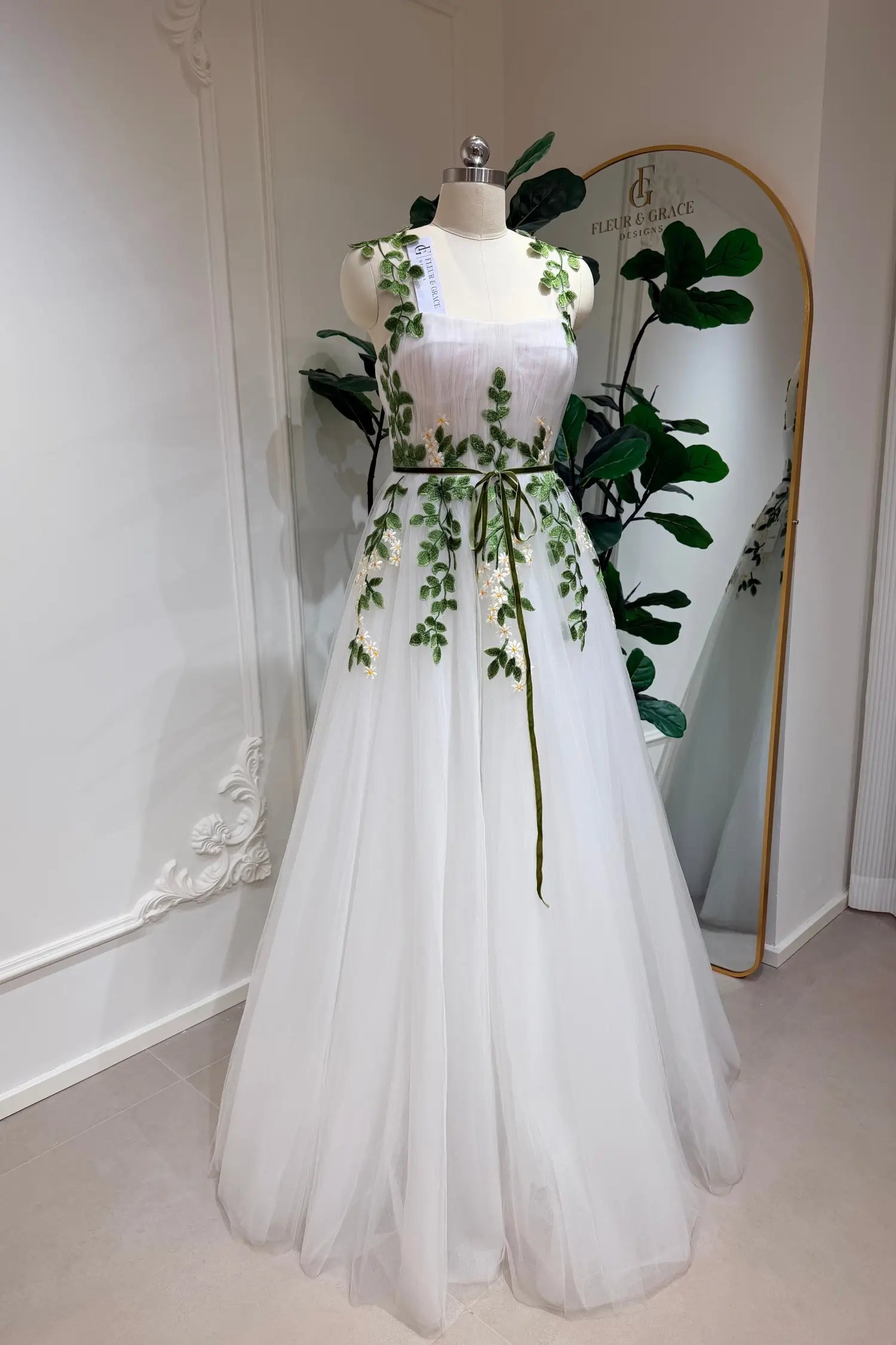 Ivory tulle A-line wedding gown featuring green leaf and daisy embroidery, a sweetheart bodice, and a thin green belt.