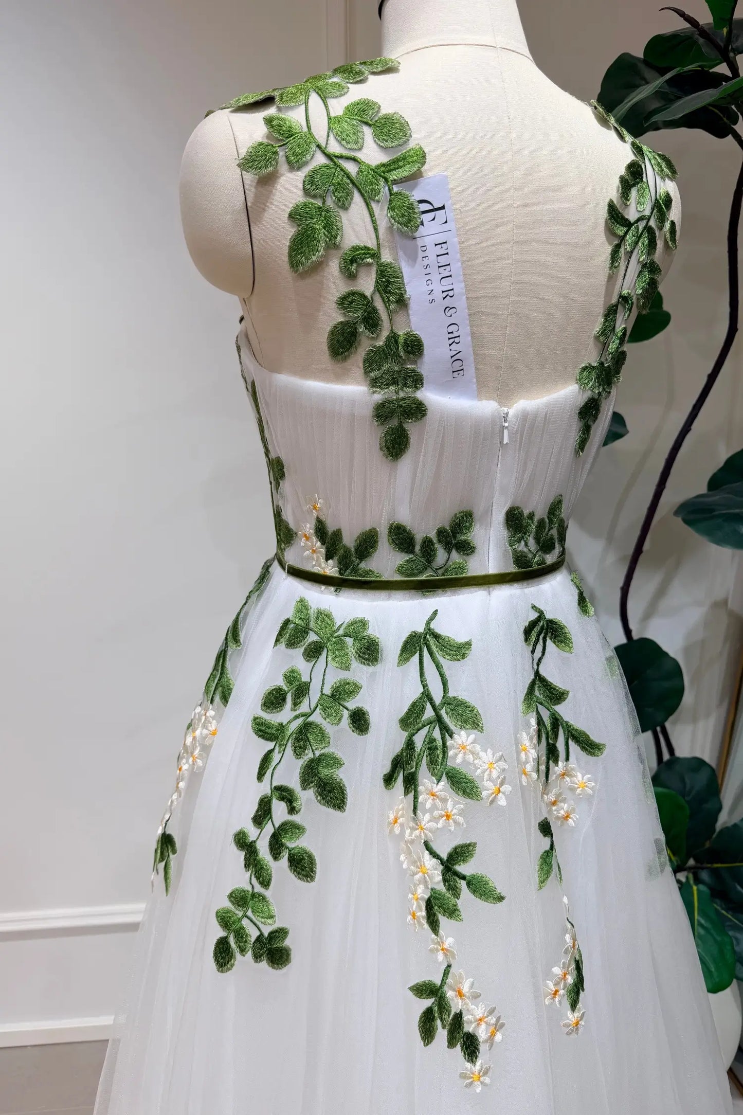 Ivory tulle wedding dress with green leaf embroidery, daisy accents, and a thin green belt on a mannequin.