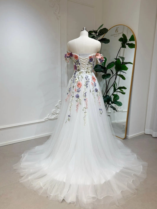 Off-shoulder A-line wedding dress with colorful 3D floral appliqué, lace-up corset back, and flowing tulle skirt.