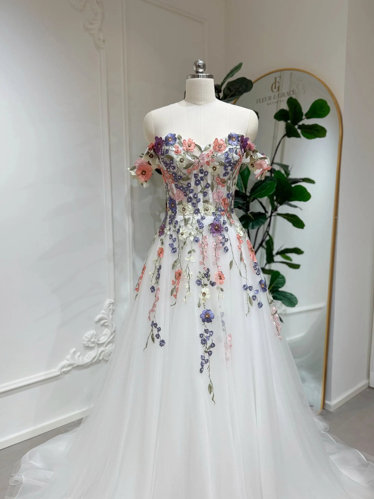 Off-shoulder A-line wedding dress with colorful 3D floral appliqué, beaded details, and airy white tulle on a mannequin.